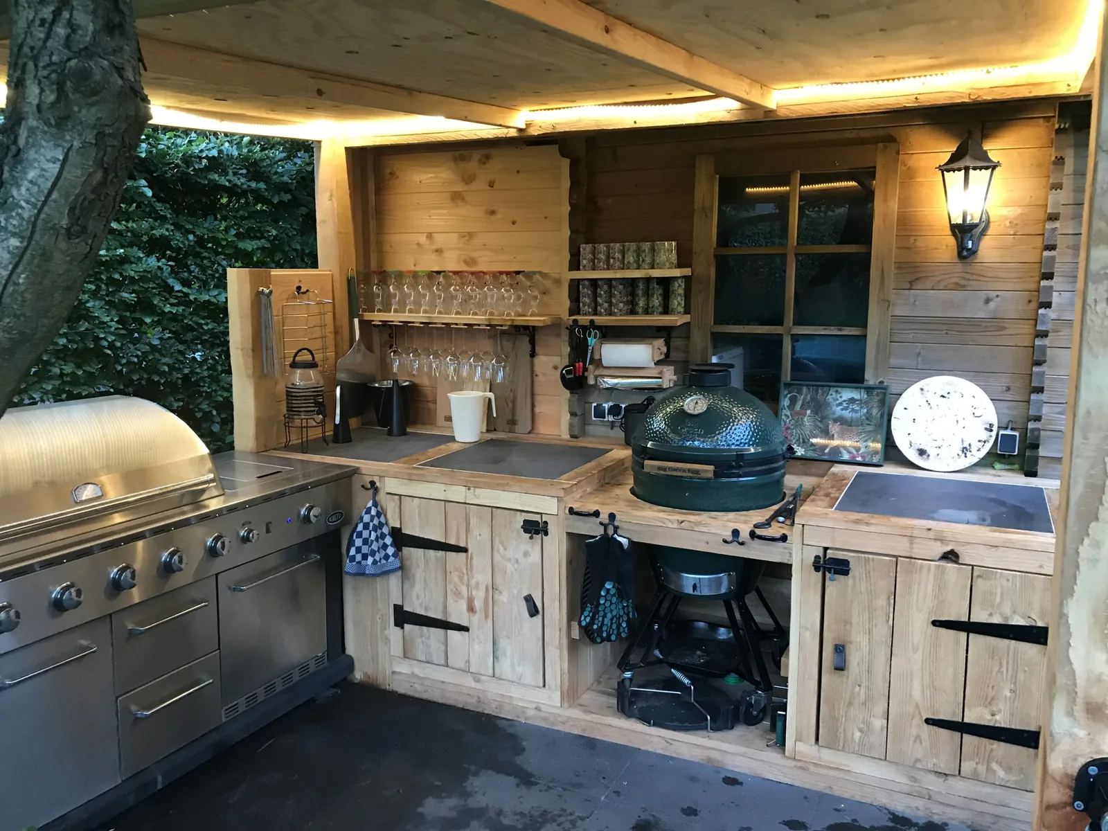 Outdoor kitchen built by Eduard Renger: custom timber cabinets with steel handles, a gas grill and a Big Green Egg under a wooden canopy